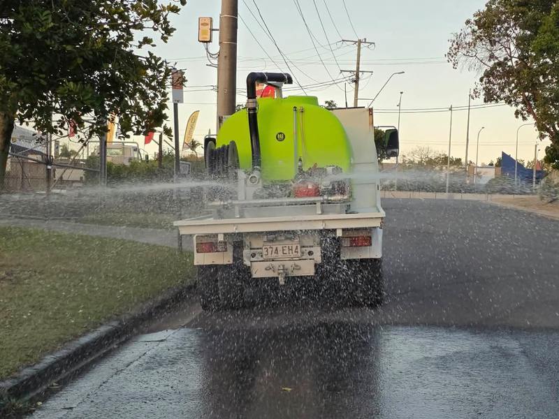 The water cart from behind, spray heads laying water across the full width of a road